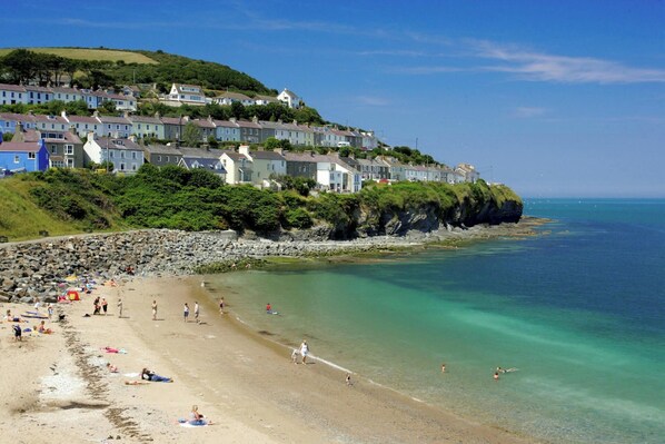 A coastal town with terraced houses, a sandy beach, and clear turquoise waters. - Cwm Llethi, New Quay (New Quay)