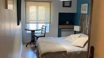 A bedroom with a bed, a desk, and a chair. There is a window with blinds, a ceiling light, and a wall-mounted picture.