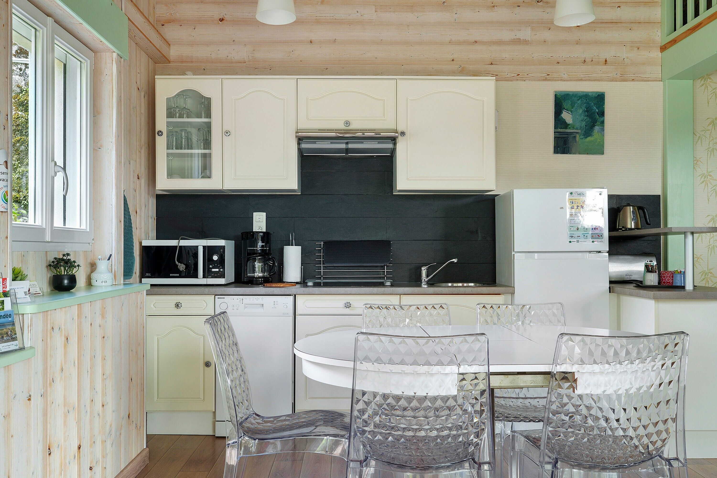 A kitchen with white cabinets, a black backsplash, a white refrigerator, a microwave, a dishwasher, and a dining table with clear chairs.