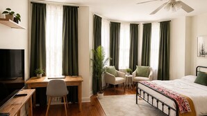 Habitación de hotel con piso de madera, una cama, un escritorio con silla, televisor y ventilador de techo.
