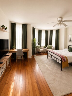 Ein Hotelzimmer mit Holzboden, einem Bett, einem Schreibtisch mit Stuhl, einem Fernseher und einem Deckenventilator.