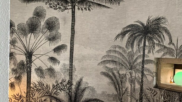 A bedroom with a bed, a wall mural of palm trees, a nightstand, and a lamp.
