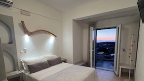 A bedroom with a bed, a bedside table, a wall-mounted light fixture, and a view through an open door to the outside. - Alexandras studios  (Irakleia)