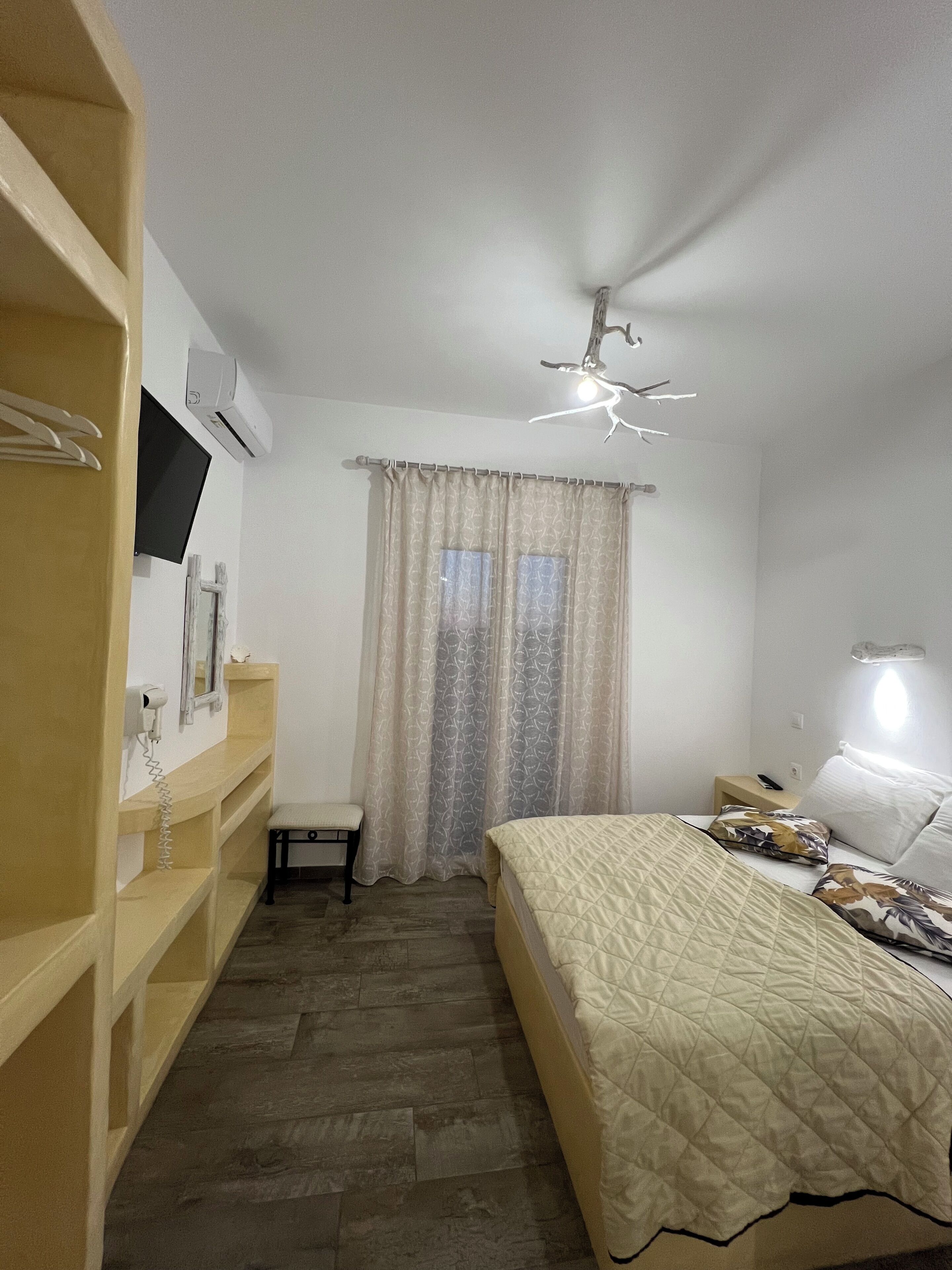 A bedroom with a bed, a bedside table, a chair, a wardrobe, a mirror, a window with curtains, and a ceiling light fixture.