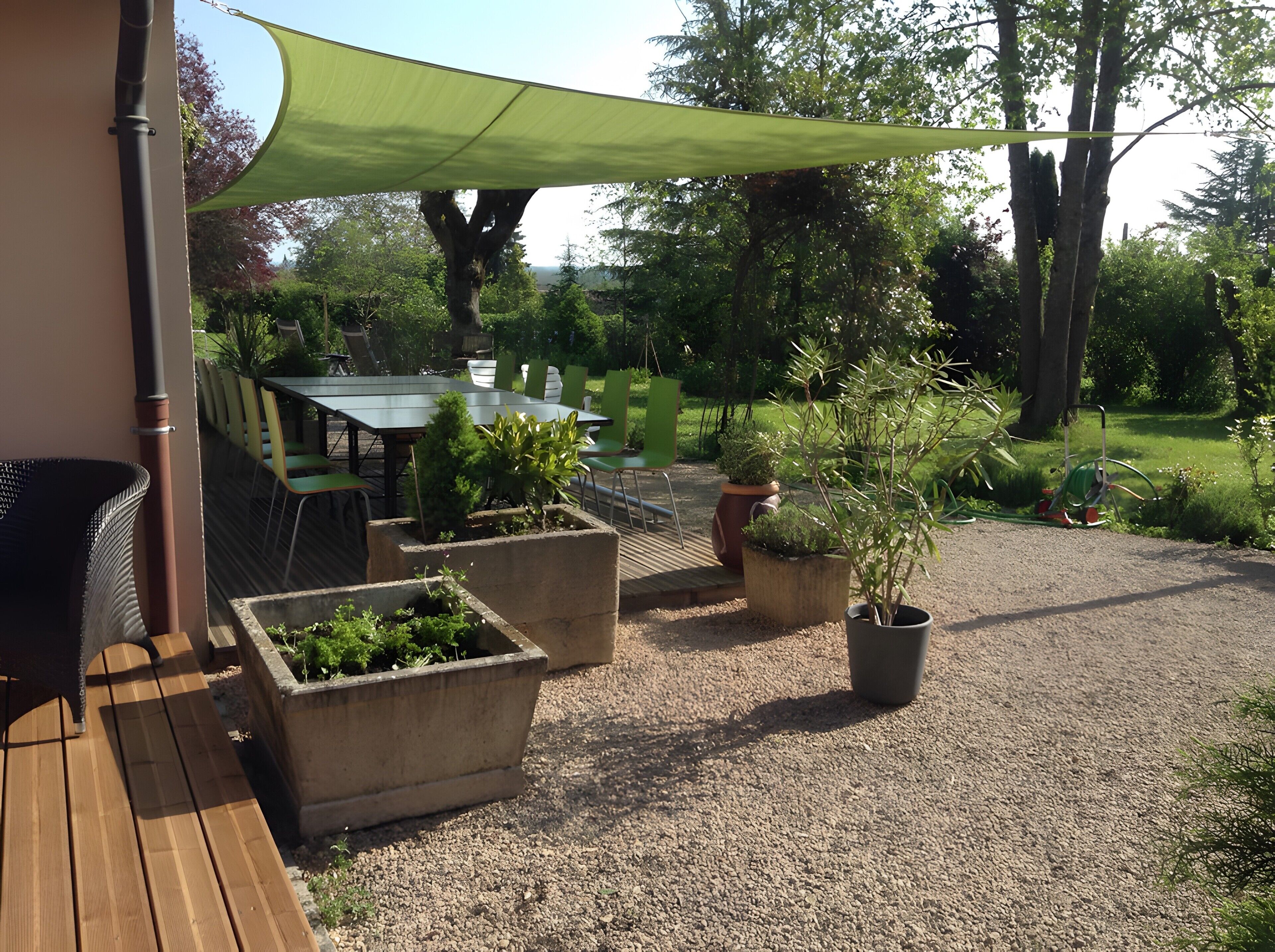 Une terrasse avec une table et des chaises, entourée de jardinières et protégée par une voile d’ombrage verte.