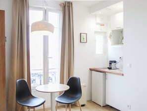 Ein Zimmer mit Esstisch, zwei Stühlen, einer Küchenzeile und einem Fenster mit Vorhängen.