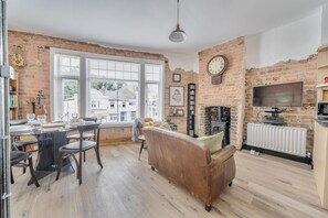 A living room with a brick wall, a fireplace, a television, and a dining area with a table and chairs. - Stylish 2Bed Flat Near Hampton Court & RiverThames - Pass the Keys (East Molesey)