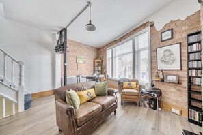 A living room with a brown leather sofa, a small round table with a speaker, a bookshelf, and a brick wall. - Stylish 2Bed Flat Near Hampton Court & RiverThames - Pass the Keys (East Molesey)