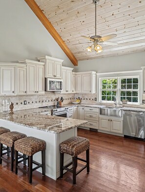 A spacious kitchen with white cabinetry, a large island with stools, a ceiling fan, and a view of greenery outside. - Riverside at Pear Tree Point--Waterfront Escape (Chestertown)