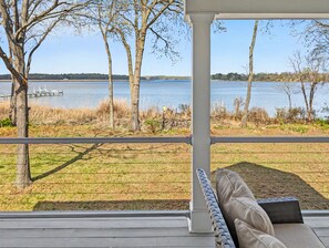 A covered outdoor area with a view of a lake, a wooden deck, and a wicker chair with cushions. - Riverside at Pear Tree Point--Waterfront Escape (Chestertown)