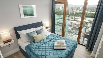 A bedroom with a large bed, a bedside table with a lamp, a framed picture on the wall, and a view of a cityscape through a window.
