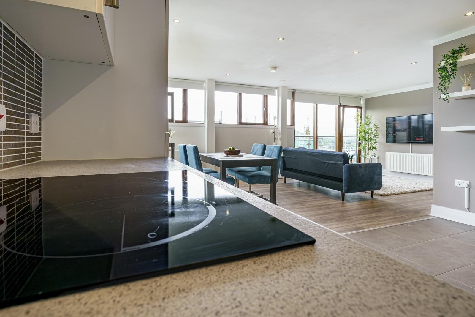 A modern kitchen with a black countertop, a dining area with a table and chairs, a sofa, and a TV mounted on the wall.