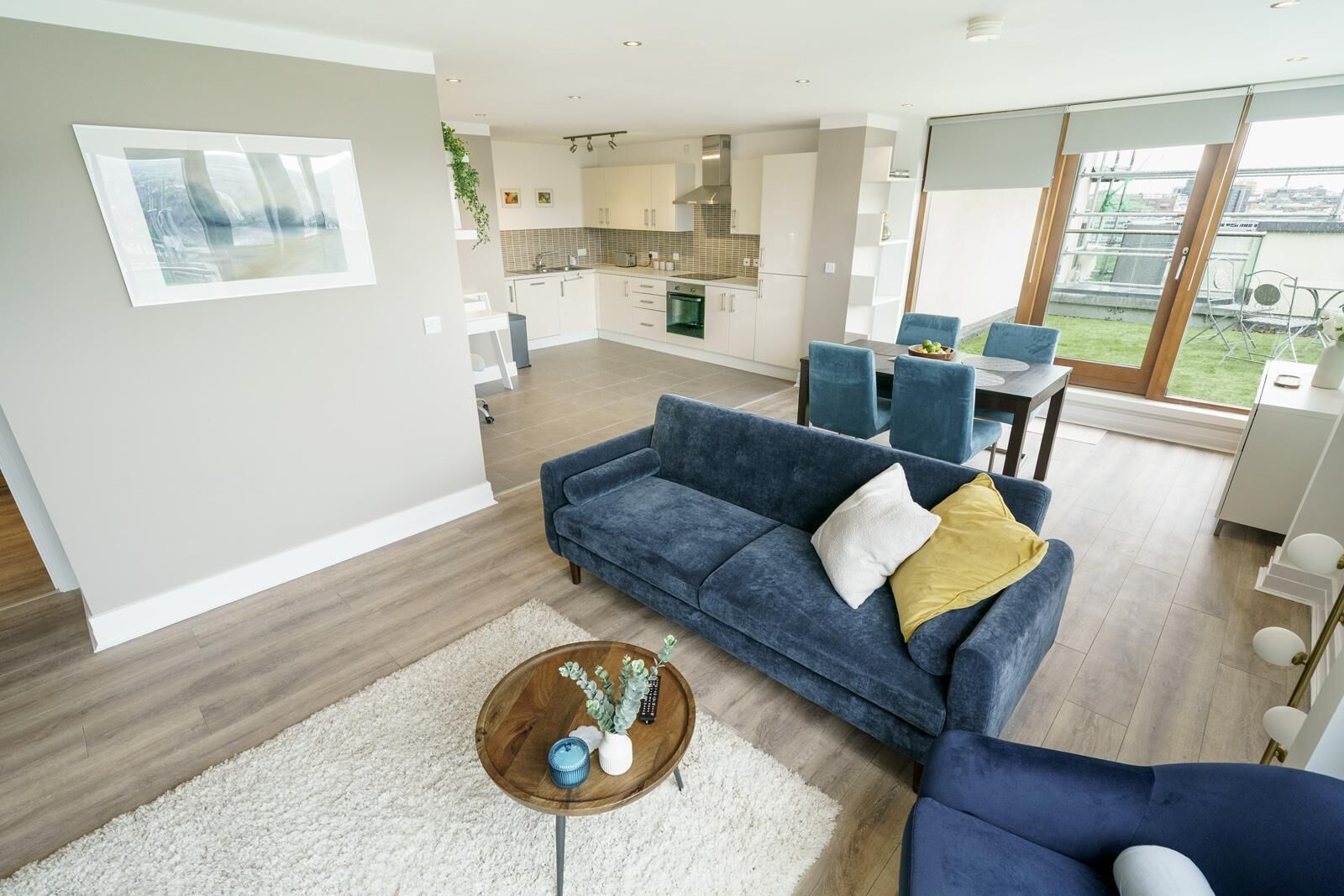 A modern living room with a blue sofa, a coffee table with a vase, and a view of the kitchen and dining area.