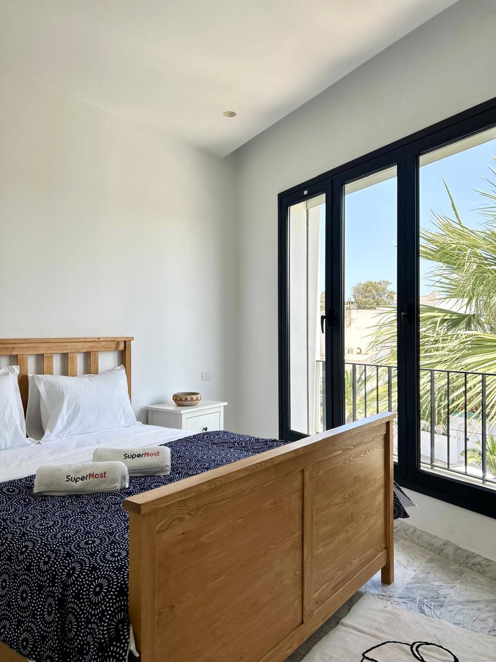 A bedroom with a wooden bed, white pillows, a nightstand, and a view of a balcony with palm trees.