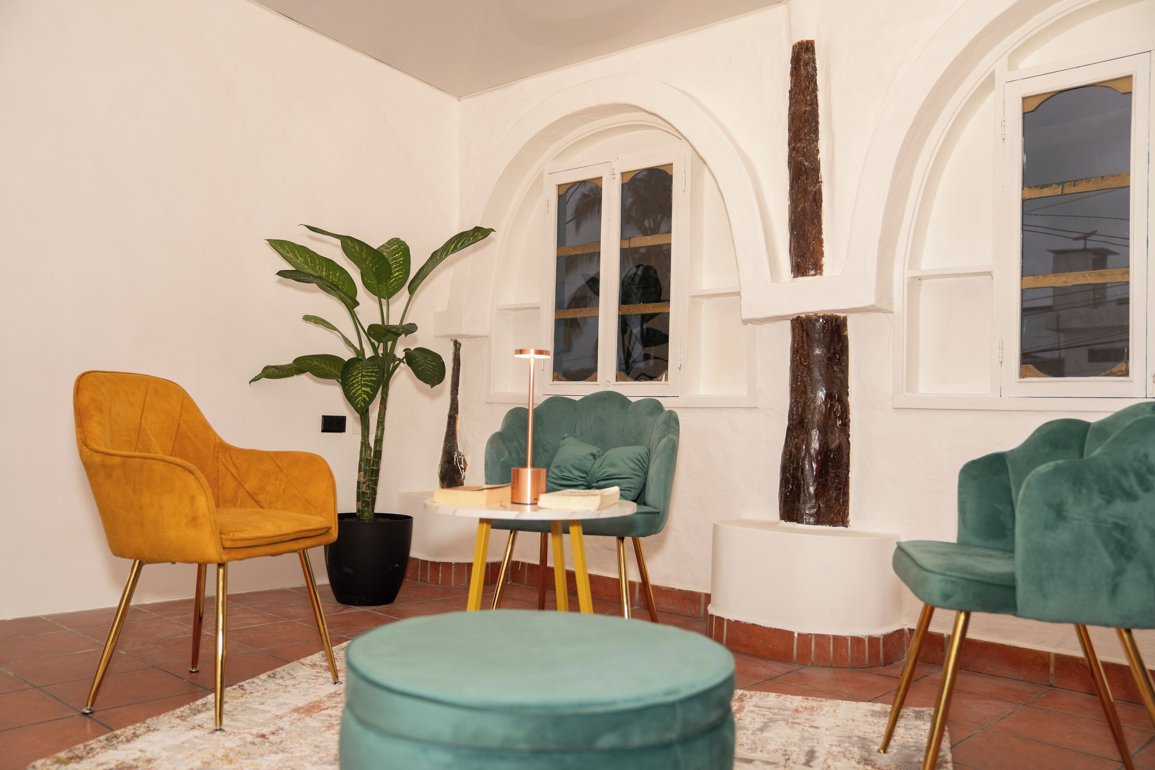 A room with a teal ottoman, a yellow chair, and a green chair with a table between them.