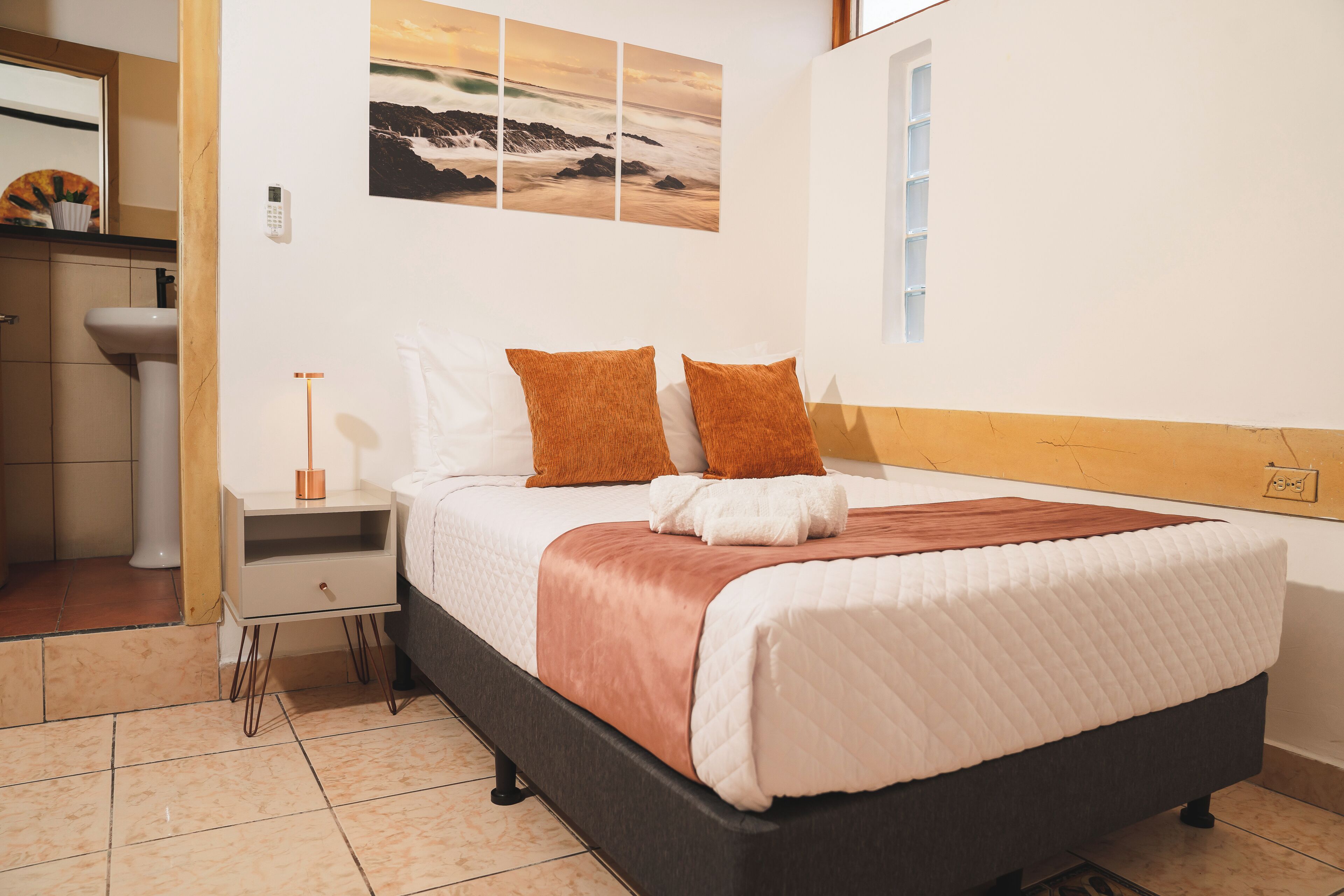 Habitación de hotel con cama, mesita de noche, ventana y un cuadro colgado en la pared.