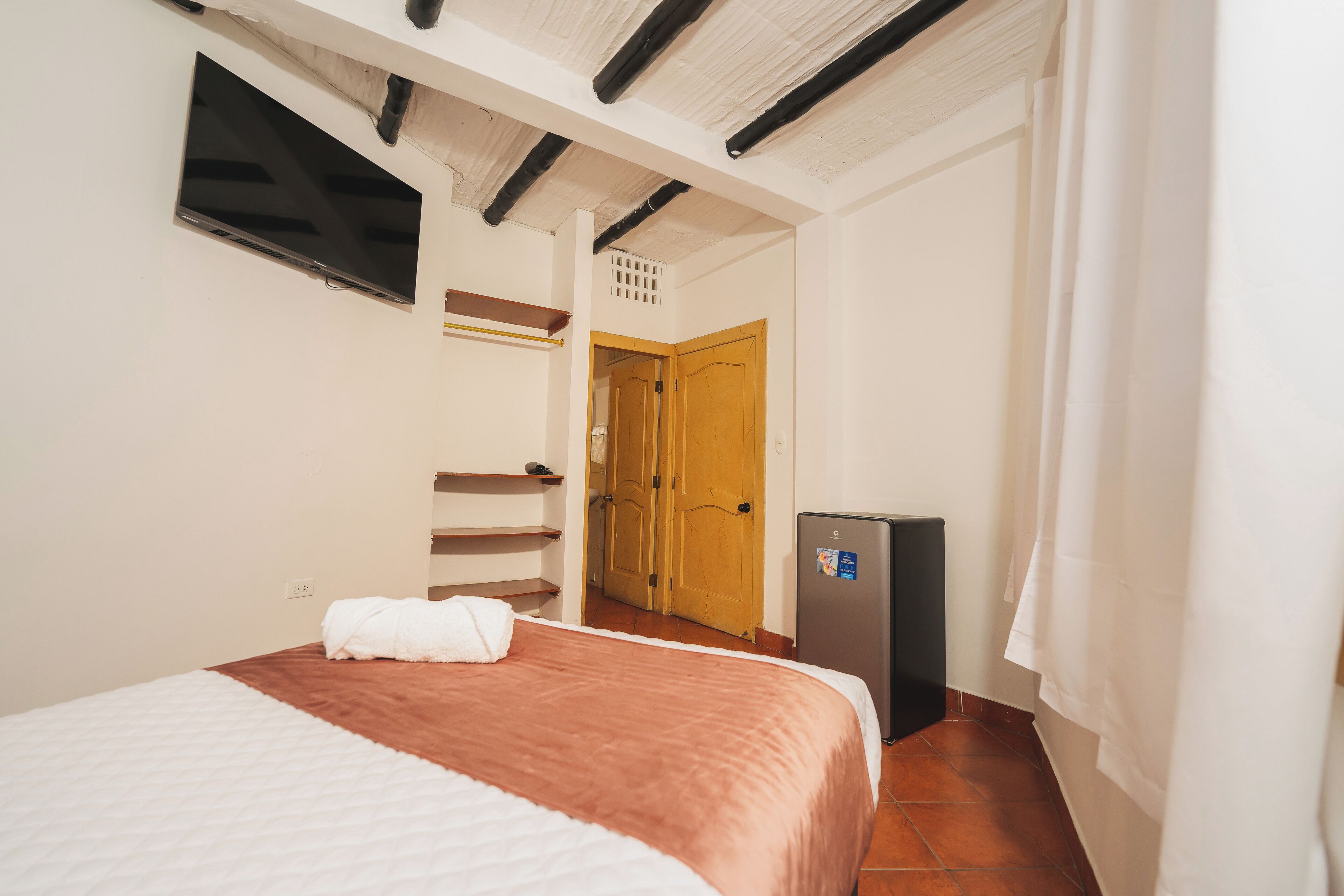 A bedroom with a bed, a television, a refrigerator, and a door leading to another room.