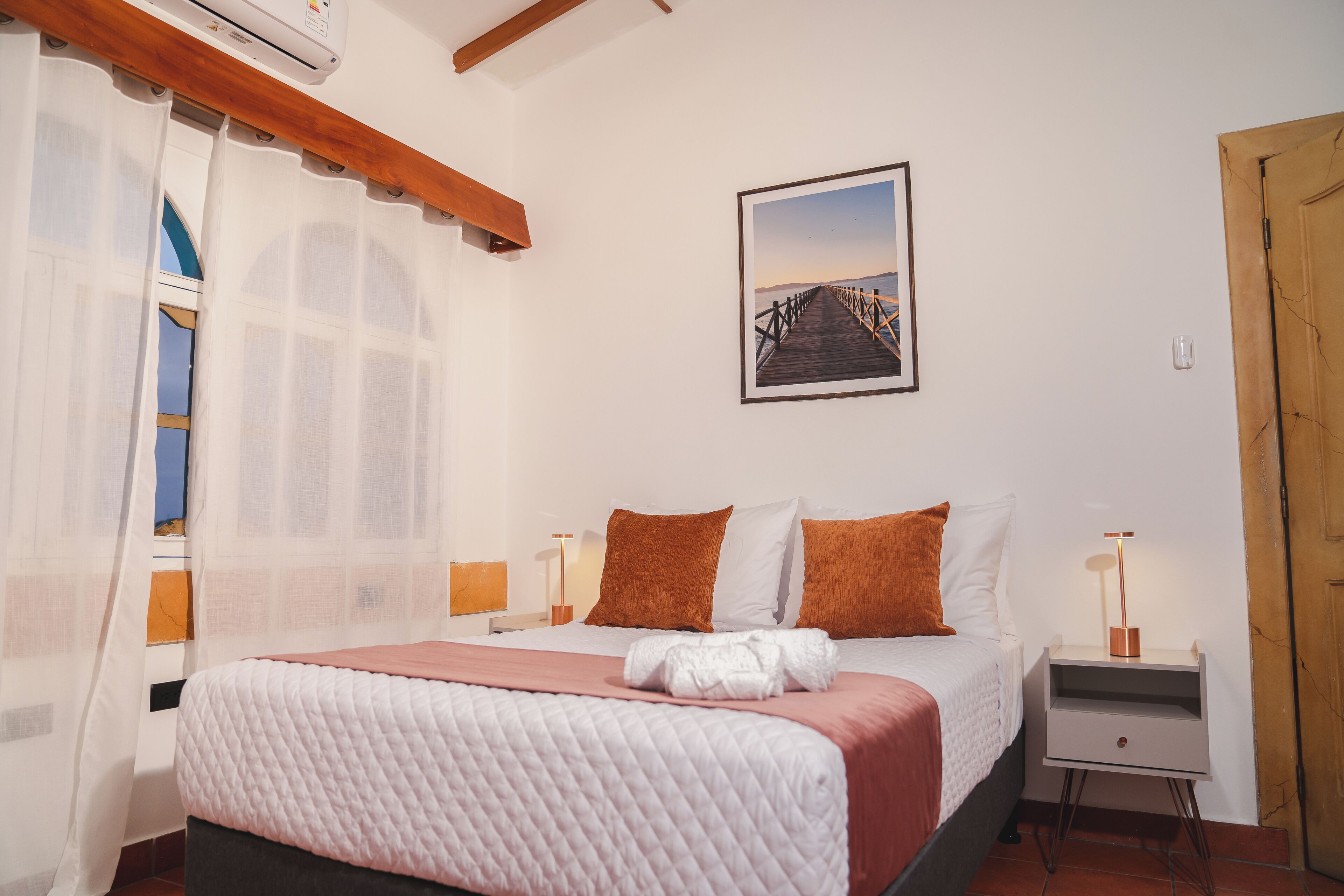 A bedroom with a bed, a nightstand, a lamp, a picture on the wall, and a window with curtains.