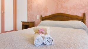 Un lit bien fait, avec des serviettes pliées et un petit vase de fleurs, une table de chevet en bois et une porte dotée d’une poignée en laiton.