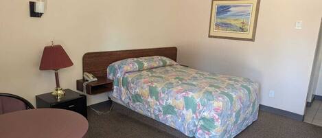 A hotel room with a bed, a desk, a chair, a lamp, a painting on the wall, and a carpeted floor.