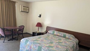 A hotel room with a bed, a desk with a chair, a lamp, and an air conditioning unit. - Algonquin Motor Lodge (Edmonton)