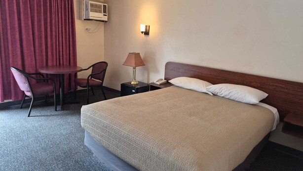A hotel room with a bed, a desk with a chair, a lamp, an air conditioning unit, and a telephone. - Algonquin Motor Lodge (Edmonton)