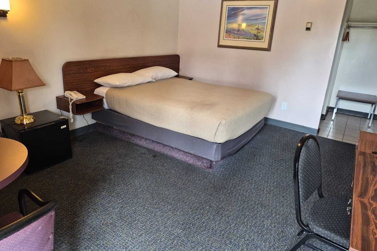 A hotel room with a bed, a desk, a chair, a lamp, a phone, and a painting on the wall.