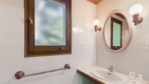Ein Badezimmer mit einem raumhohen Holzrahmenfenster, einem runden Spiegel mit Wandleuchten, einem weißen Waschbecken und einer Haltestange.