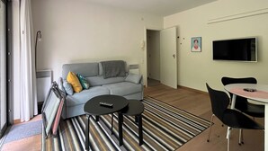 Ein Wohnzimmer mit grauem Sofa, zwei schwarzen Tischen, einem Streifen Teppich, einem Fernseher an der Wand und einer Tür zu einem anderen Raum.