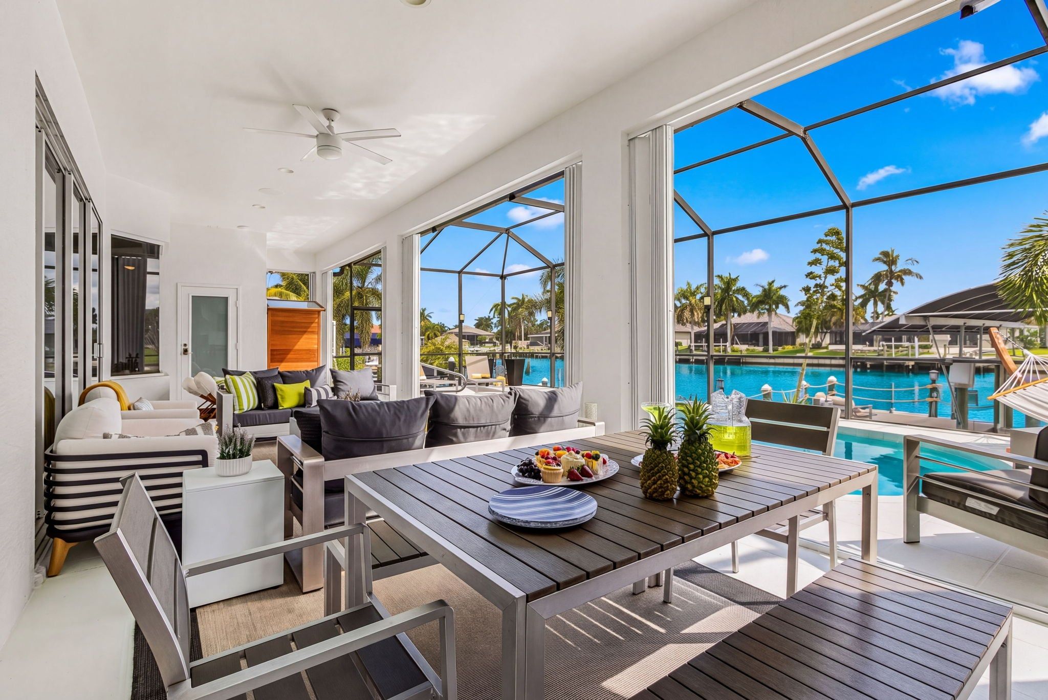 A modern outdoor seating area with a pool view, a dining table with fruit, and a ceiling fan.
