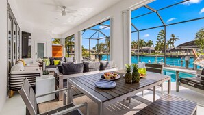 A modern indoor space with a pool view, a dining area with a table set for two, and a seating area with a sofa and chairs. - Heated Pool, Gulf Access & Waterfront Luxury Escape - Seaside Social - Roelens (Cape Coral)