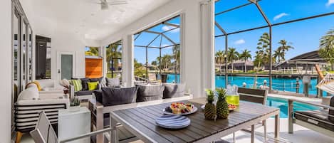 A modern outdoor seating area with a pool view, a dining table with fruit, and a ceiling fan.