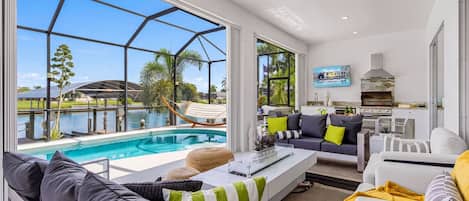 Un salon moderne avec vue sur la piscine, un ensemble de canapés, un coin repas et une cuisine équipée d’un barbecue.