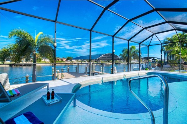 Ein überdachter Innenpool, eine Lounge mit Tisch und Stühlen sowie Blick auf ein Gewässer und Palmen.