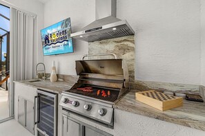 An outdoor kitchen with a grill, a television displaying 'Roelens Vacations', and a chessboard. - Heated Pool, Gulf Access & Waterfront Luxury Escape - Seaside Social - Roelens (Cape Coral)