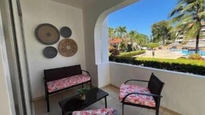 A balcony with a sofa and chairs, decorative plates on the wall, and a view of a pool and greenery. - 1 bedroom and 1 bathroom apartment in Monte Vista, Sosúa (Sosúa)
