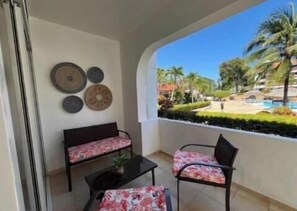 Ein Balkon mit einer Couch und Stühlen, dekorativen Tellern an der Wand sowie Blick auf einen Pool und Grünflächen.