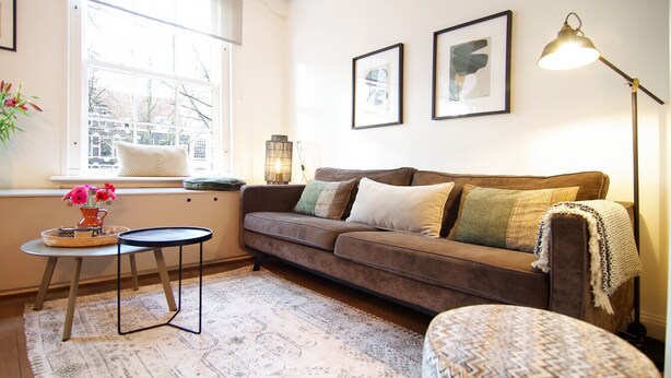 A cozy living room with a sofa, a small round table, a floor lamp, and a window with a view. - Sonnenberg - Canal side & view - Private & Central (Amsterdam)
