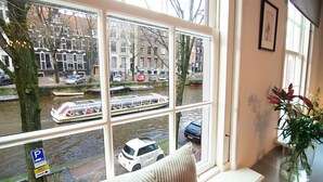 A window with a view of a canal, a white car, and a building with a blue sign. - Sonnenberg - Canal side & view - Private & Central (Amsterdam)