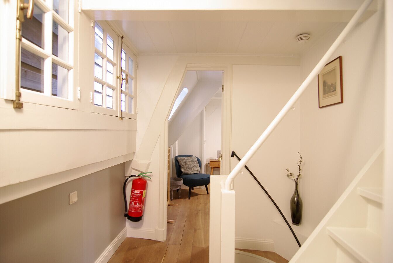 A narrow hallway with a fire extinguisher, a chair, and a vase on the wall.