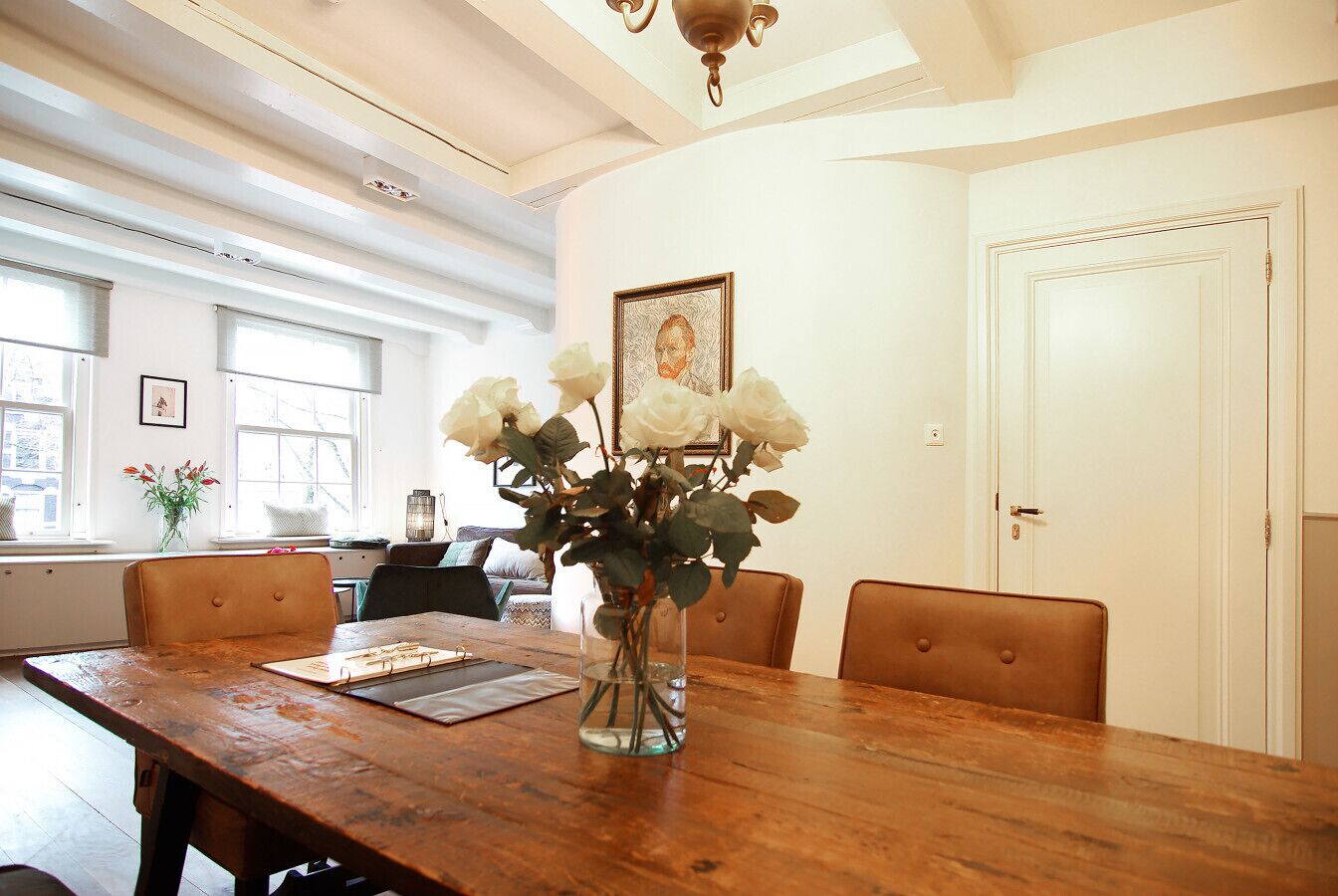 Een eetgedeelte met een houten tafel, stoelen, een vaas met bloemen en een ingelijste foto aan de muur.