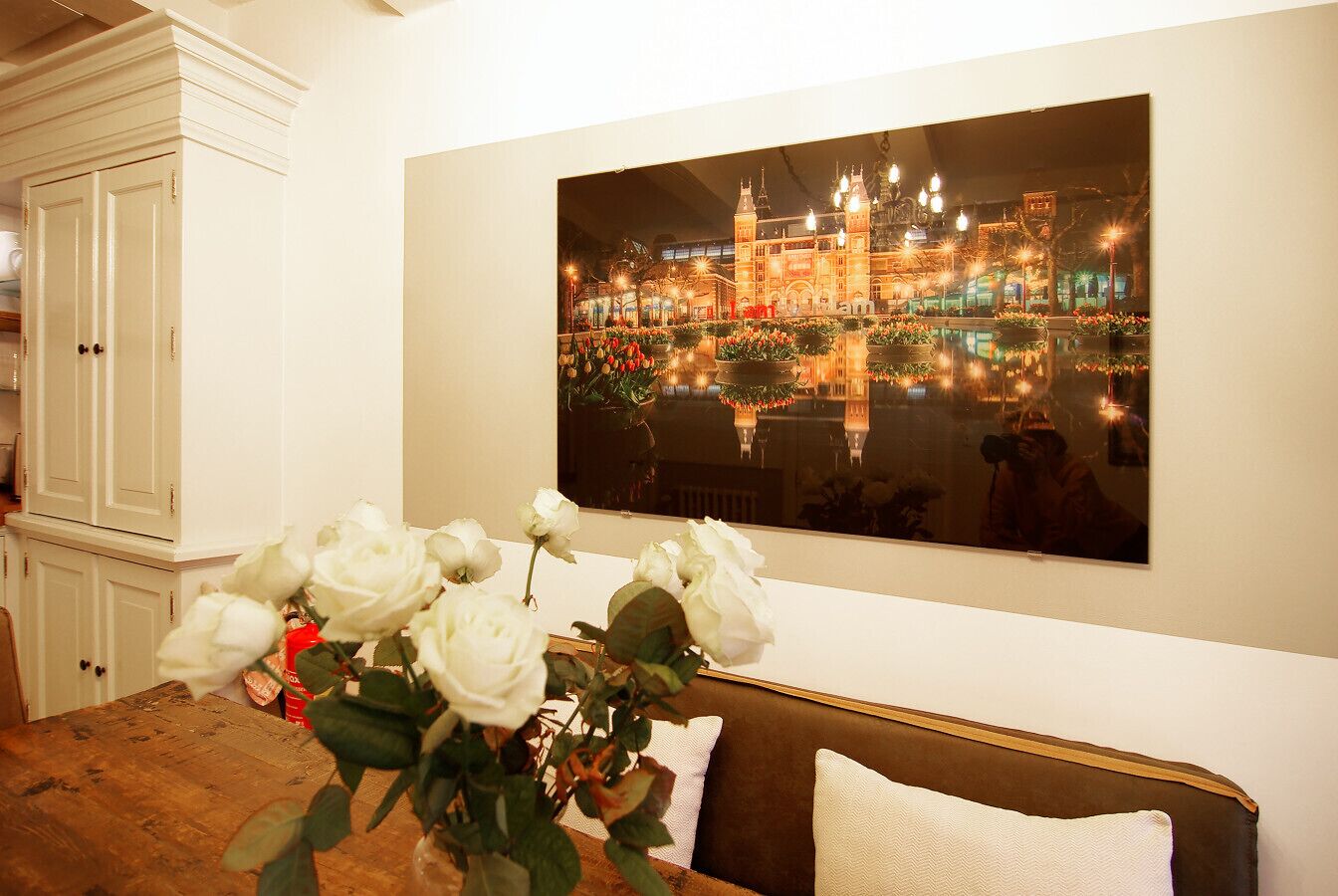 Een kamer met een groot schilderij van een stadsgezicht, een houten eettafel met een vaas witte bloemen en een bruine bank met een wit kussen.