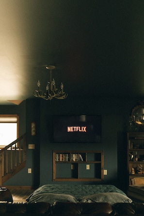 Een kamer in gedempt licht, met een bed, een boekenplank, een televisietoestel waarop 'NETFLIX' staat, en een kroonluchter.