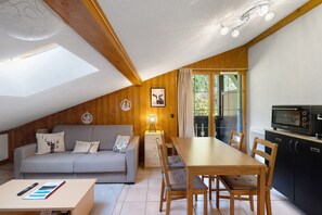 A cozy room with a sloped ceiling, wooden walls, a grey sofa with patterned cushions, a wooden dining table with chairs, a small lamp, and a kitchen area with a microwave. - Apartment 'Les Contamines Montjoie' with Mountain View, Private Terrace and Balcony (Les Contamines-Montjoie)