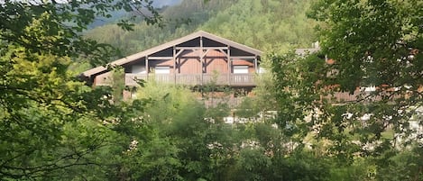 A wooden cabin with a balcony, surrounded by dense greenery and a river.
