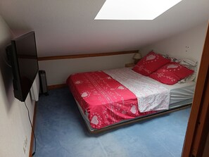 Un llit individual amb un edredó vermell i blanc, una petita taula de nit amb una làmpada, i un televisor fixat a la paret.