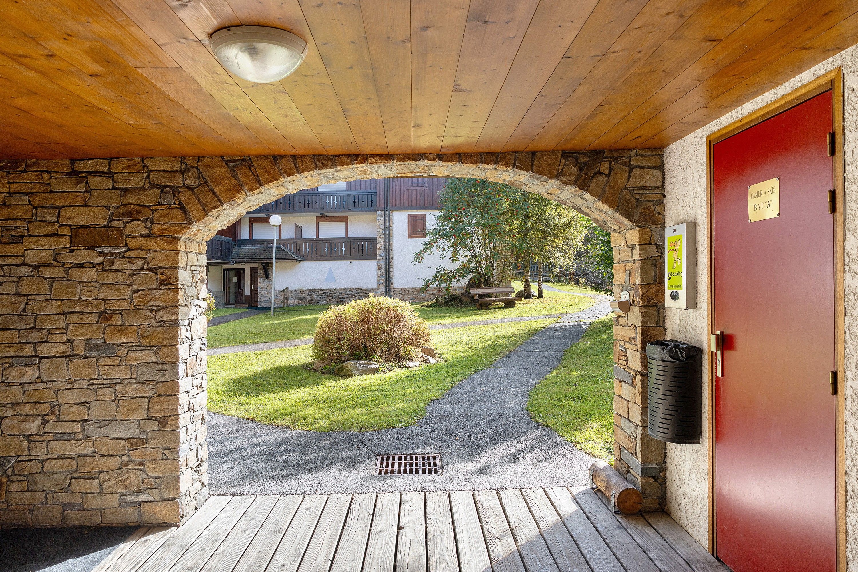 A wooden archway leading to a red door, a stone wall, a small bush, and a paved path.