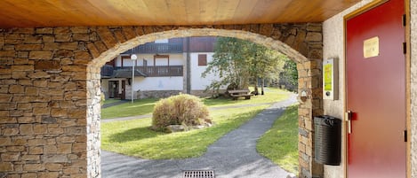 A wooden archway leading to a red door, a stone wall, a small bush, and a paved path.