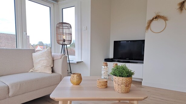 A living room with a sofa, a coffee table with decorative items, a television, and a large window. - Bright 3-bedroom Flat by the Beach (Koksijde)