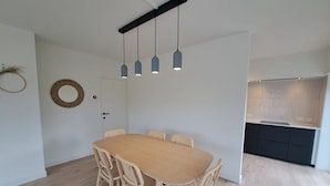 A dining area with a wooden table and wicker chairs, a white door, and a kitchen with dark cabinets. - Bright 3-bedroom Flat by the Beach (Koksijde)